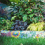 Detail of a mosaic wall and colorful plantings raised above the lawn in a small backyard.